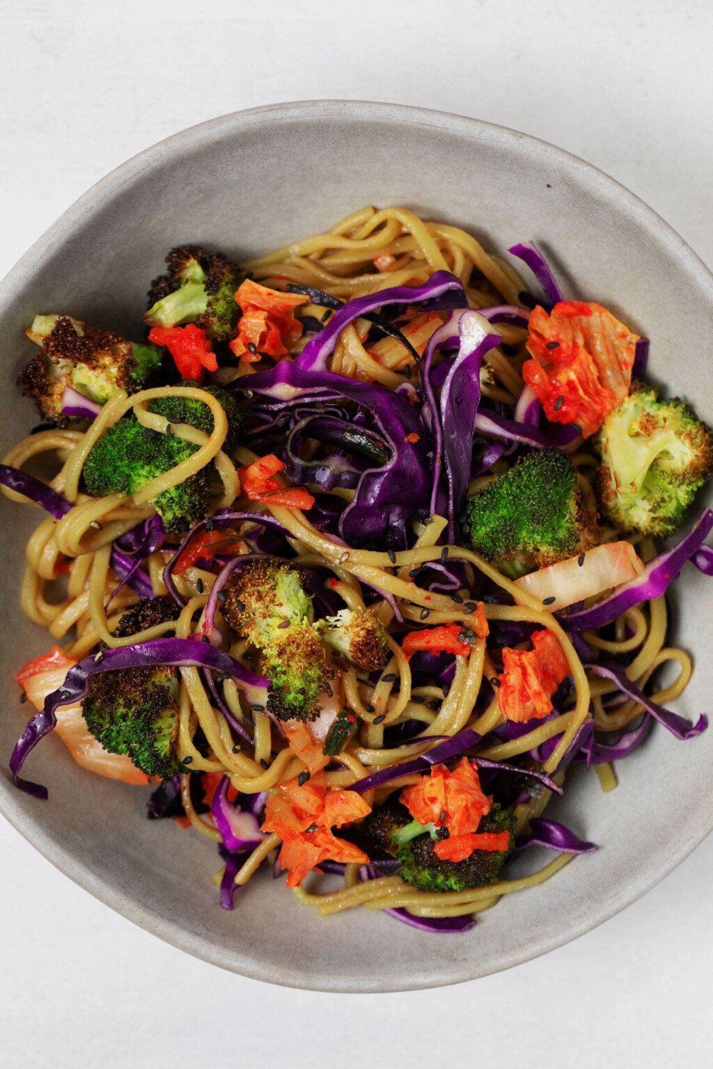 A dish of roasted broccoli kimchi noodles has been piled into a gray, ceramic bowl. The noodles also contain bright red cabbage.