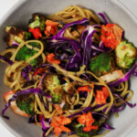 A dish of roasted broccoli kimchi noodles has been piled into a gray, ceramic bowl. The noodles also contain bright red cabbage.