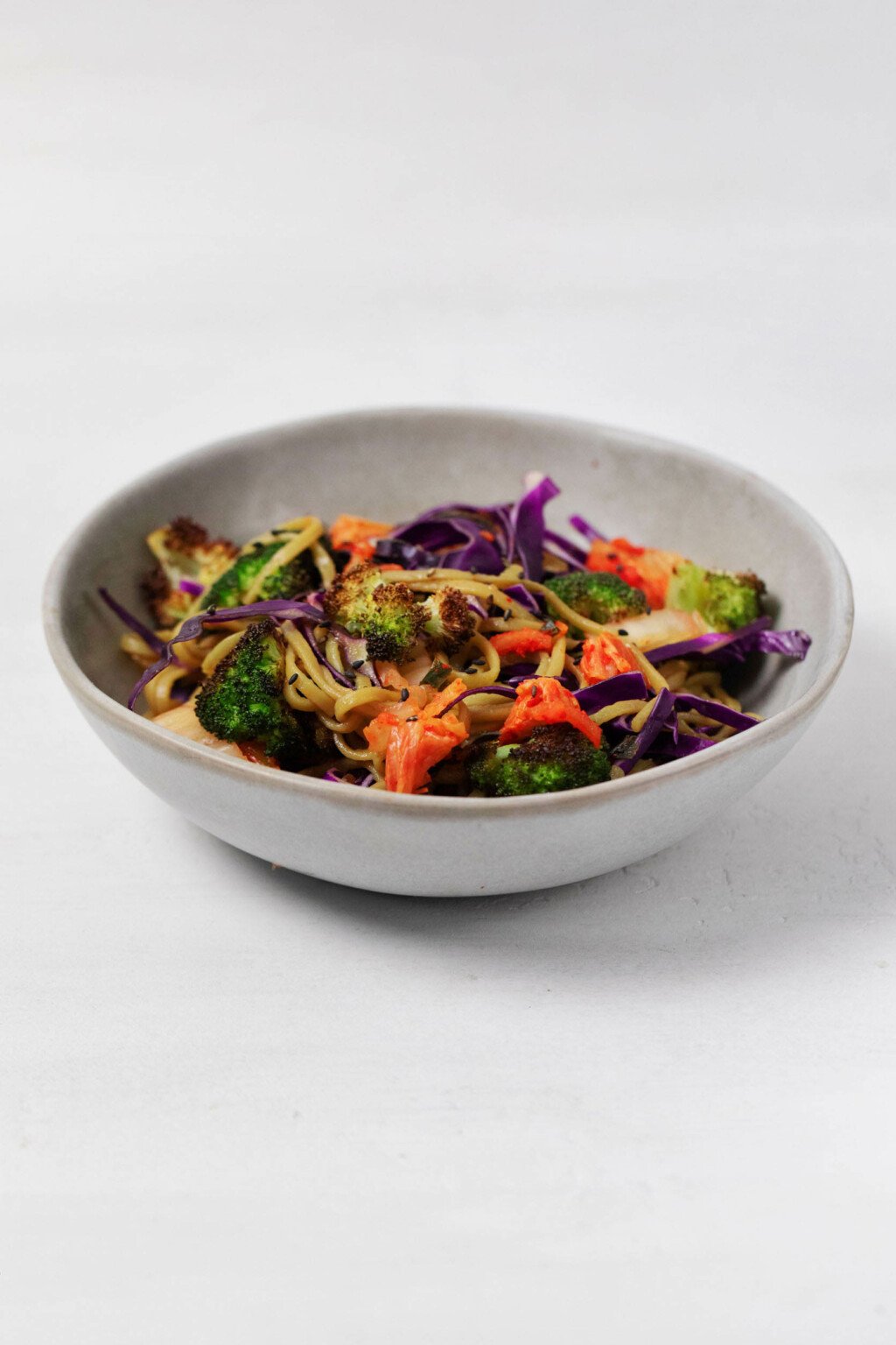 A gray ceramic bowl holds a plant based soba and vegetable dish, which is resting on a bright white surface.