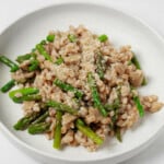 A round, rimmed white plate holds a vegan barley risotto with mushrooms and bright green asparagus pieces.