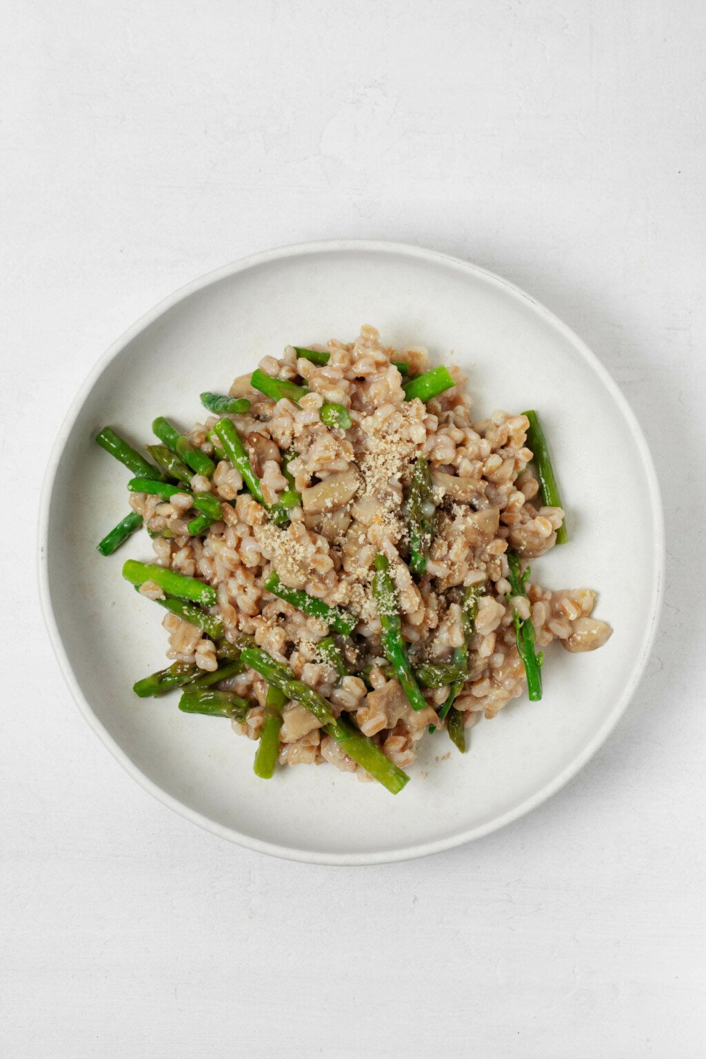 A round, rimmed white plate holds a vegan barley risotto with mushrooms and bright green asparagus pieces.