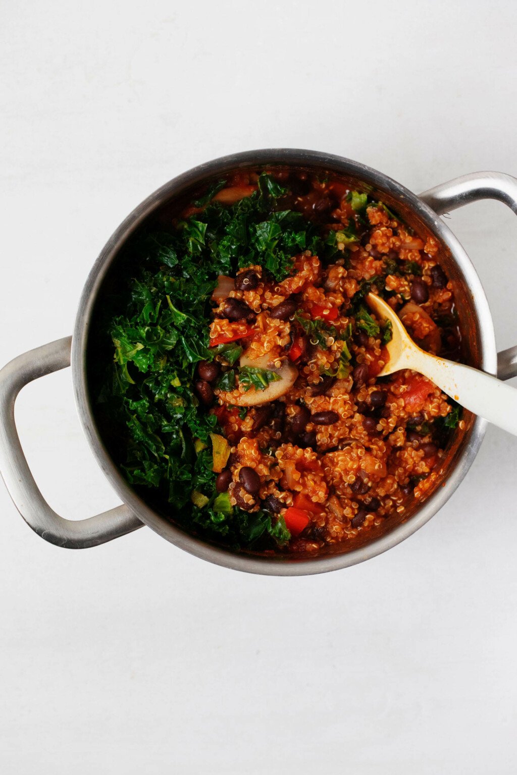 A large pot of quinoa, mushrooms, and beans is being combined with curly kale.