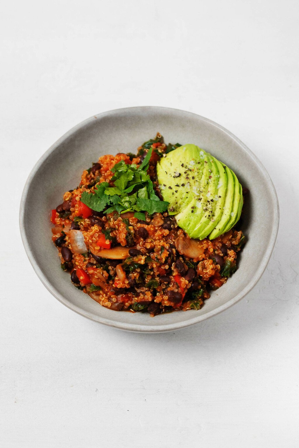 Vegan quinoa black bean chili is served in a round, white dish. It's topped with slices of avocado.