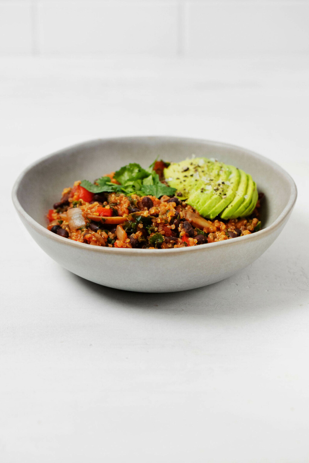 A light gray bowl holds a quinoa black bean chili. It's garnished with avocado slices and chopped herbs.