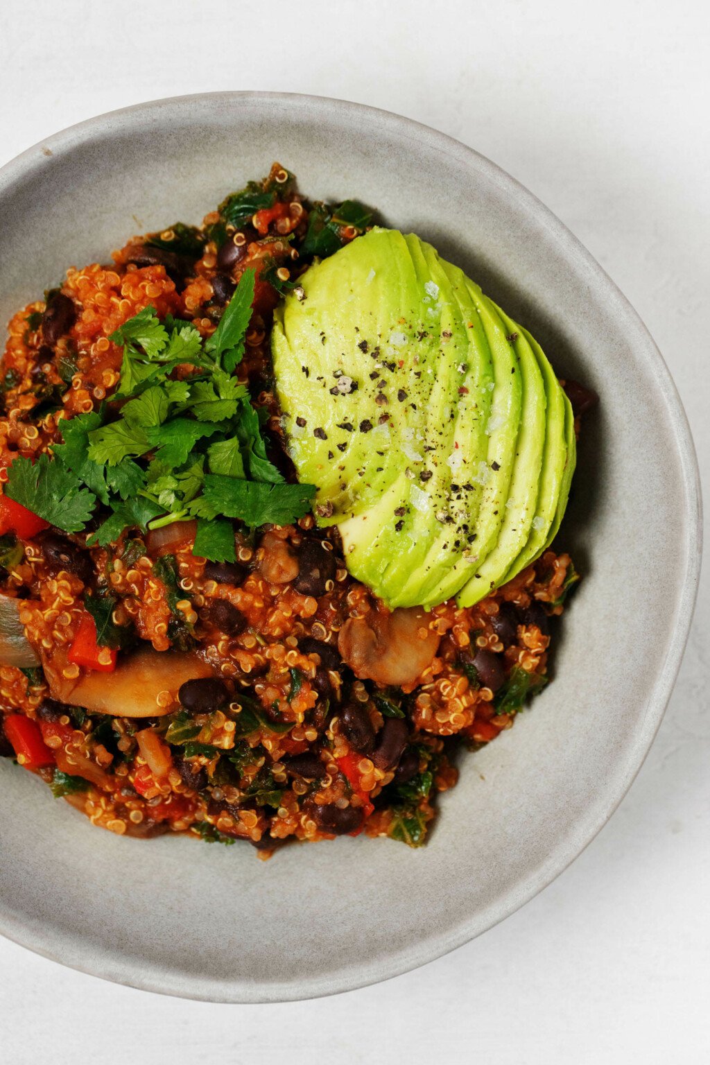 A vegan quinoa black bean chili has been served in a round bowl, resting on a white surface. It's topped with herbs and avocado.
