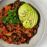 A vegan quinoa black bean chili has been served in a round bowl, resting on a white surface. It's topped with herbs and avocado.