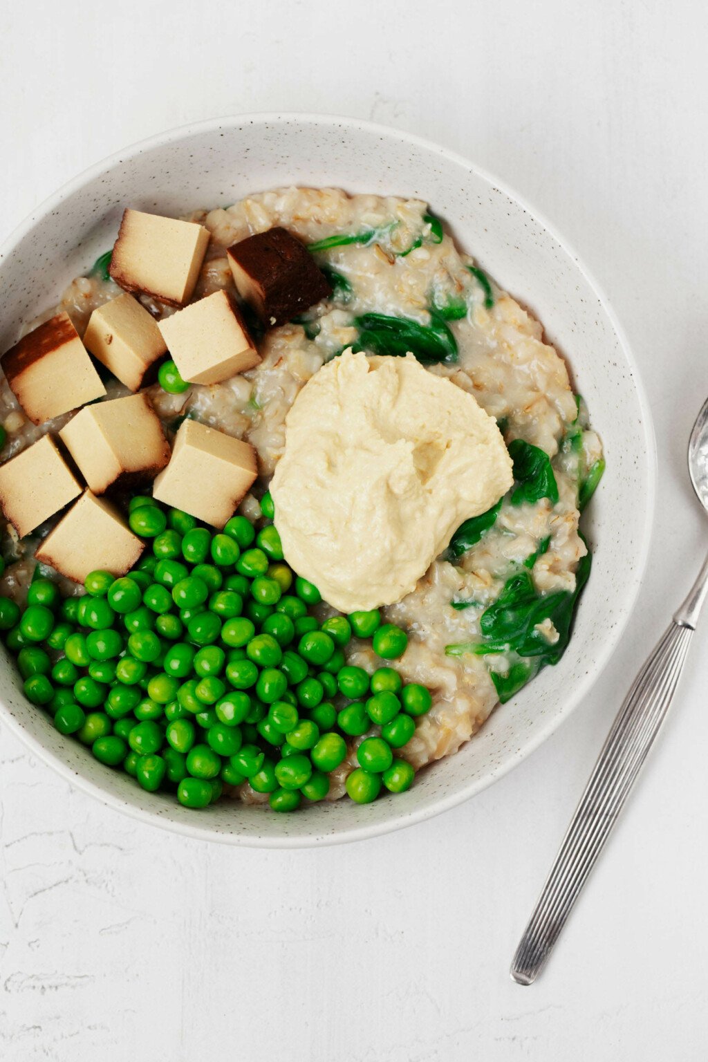 A round, white bowl is filled with savory oatmeal. The oatmeal is topped with green peas, hummus, and cubes of baked smoky tofu.