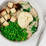 A round, white bowl is filled with savory oatmeal. The oatmeal is topped with green peas, hummus, and cubes of baked smoky tofu.