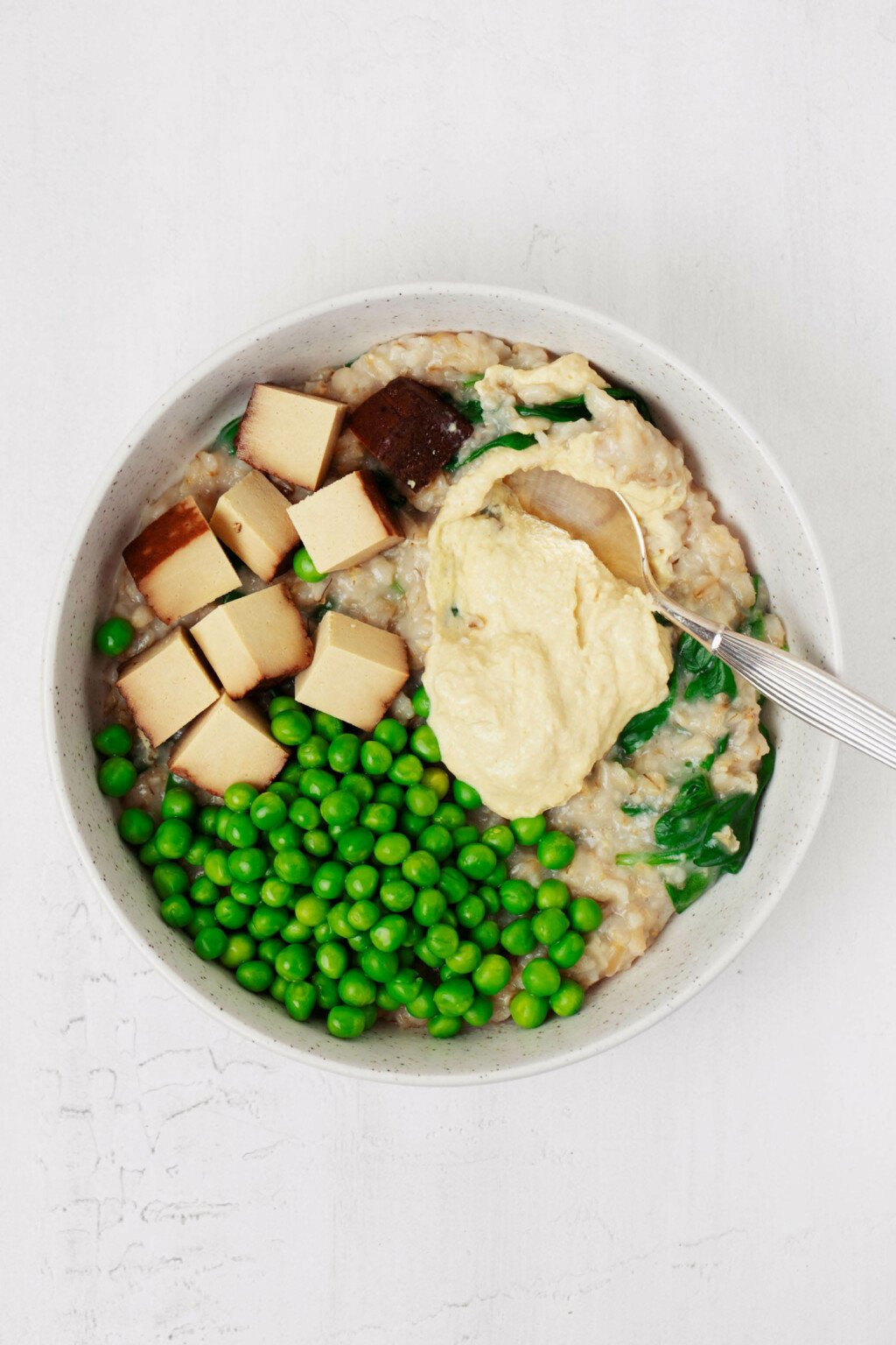 A round, white bowl rests on a white surface. The bowl is filled with plant-based ingredients and topped with a scoop of hummus.