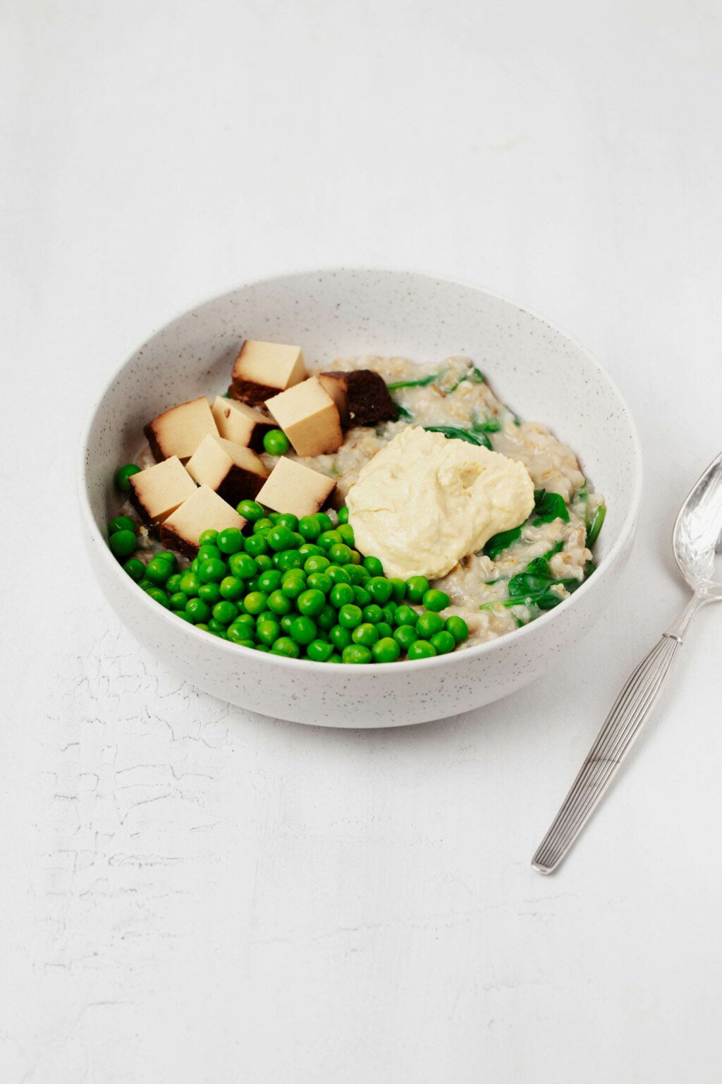 A round white bowl, containing savory oatmeal and green vegetables, is resting on a white surface. A metal spoon is perched nearby.
