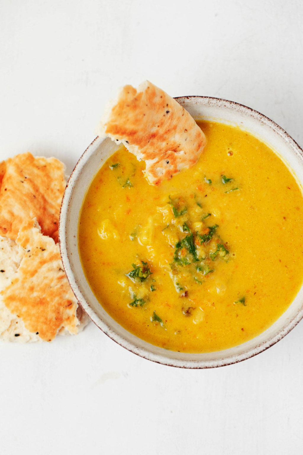 A close-up, overhead image of a bright golden-colored soup that is flecked with greens. It's served with flatbread.