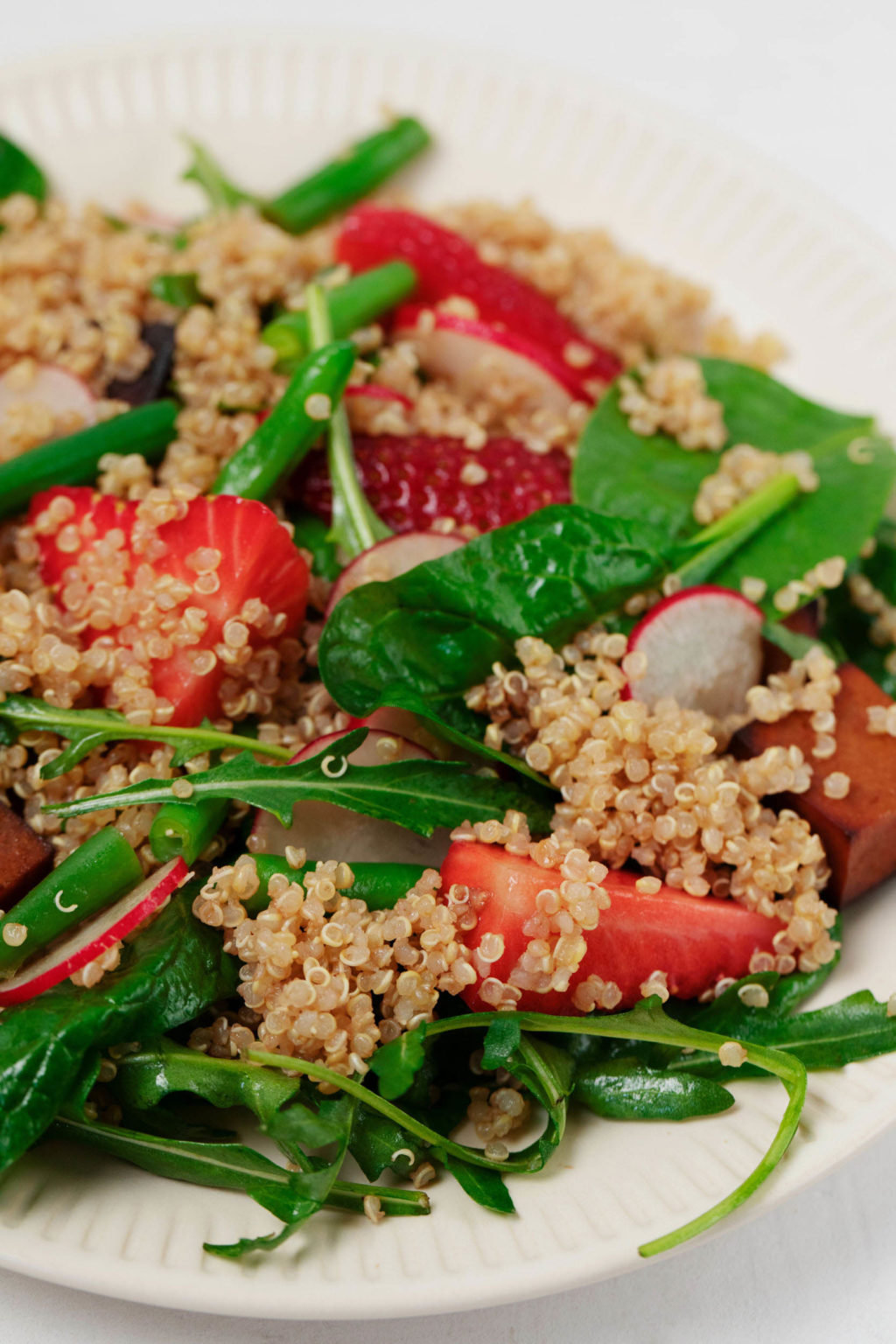 A close-up image of spinach leaves, radishes, cooked grains, strawberries, and spinach, all arranged on a white serving plate.