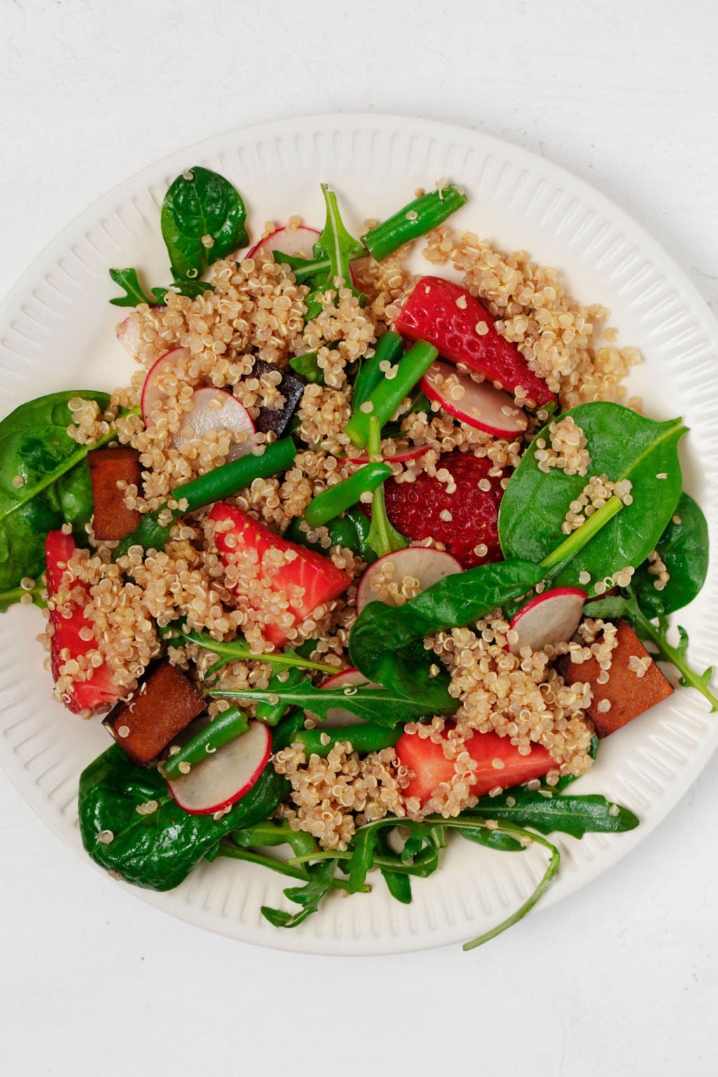 A salad of balsamic tofu, quinoa, and strawberries is arranged on a plate. Green spinach is the base of the salad. 