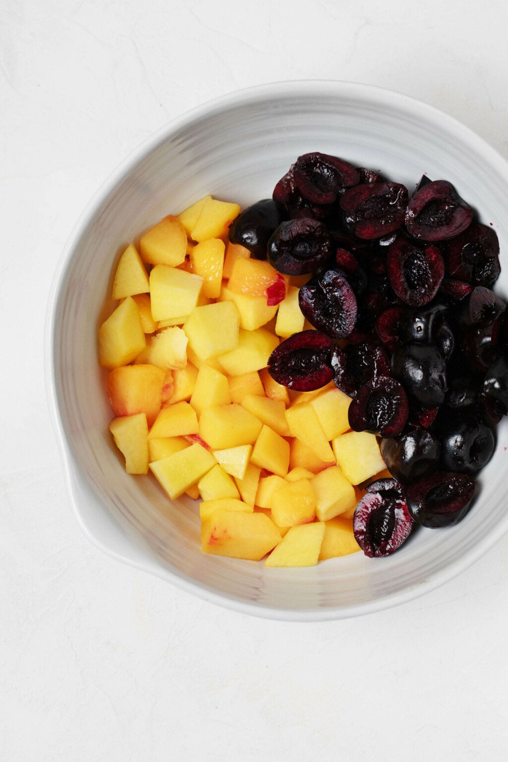 A large, white mixing bowl is filled with chopped yellow peaches and dark, sweet red cherries.