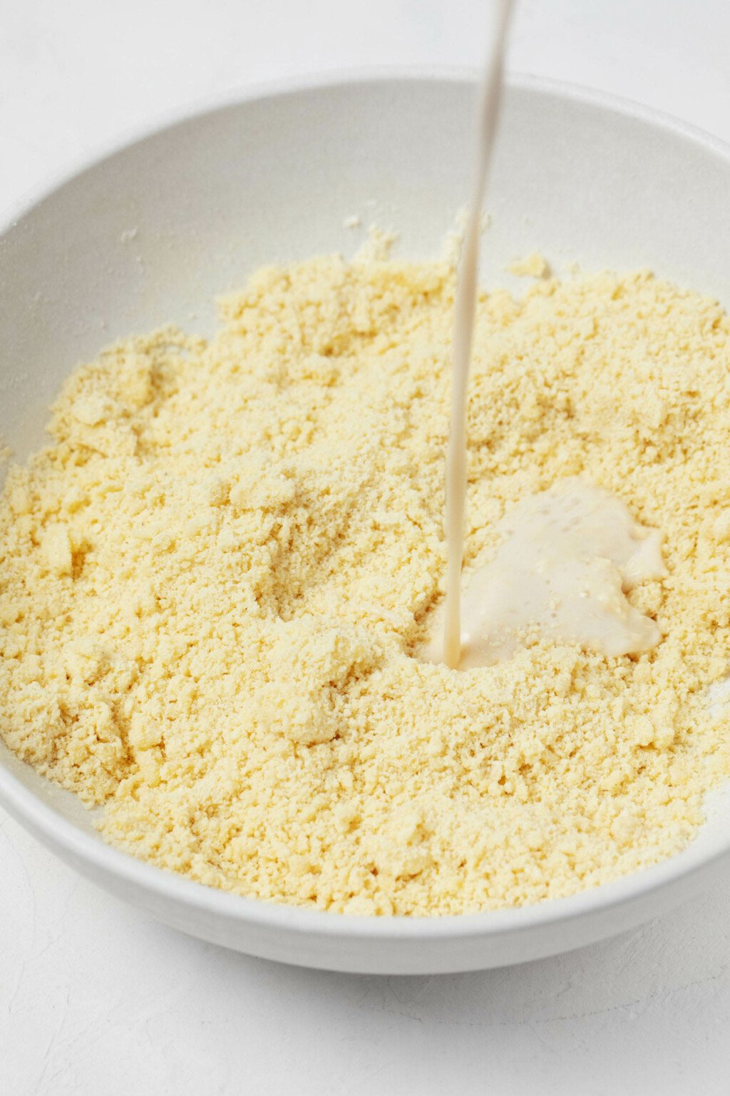 A thin stream of milk is pictured. It's being poured into a mixing bowl with dry ingredients that include cornmeal and flour.