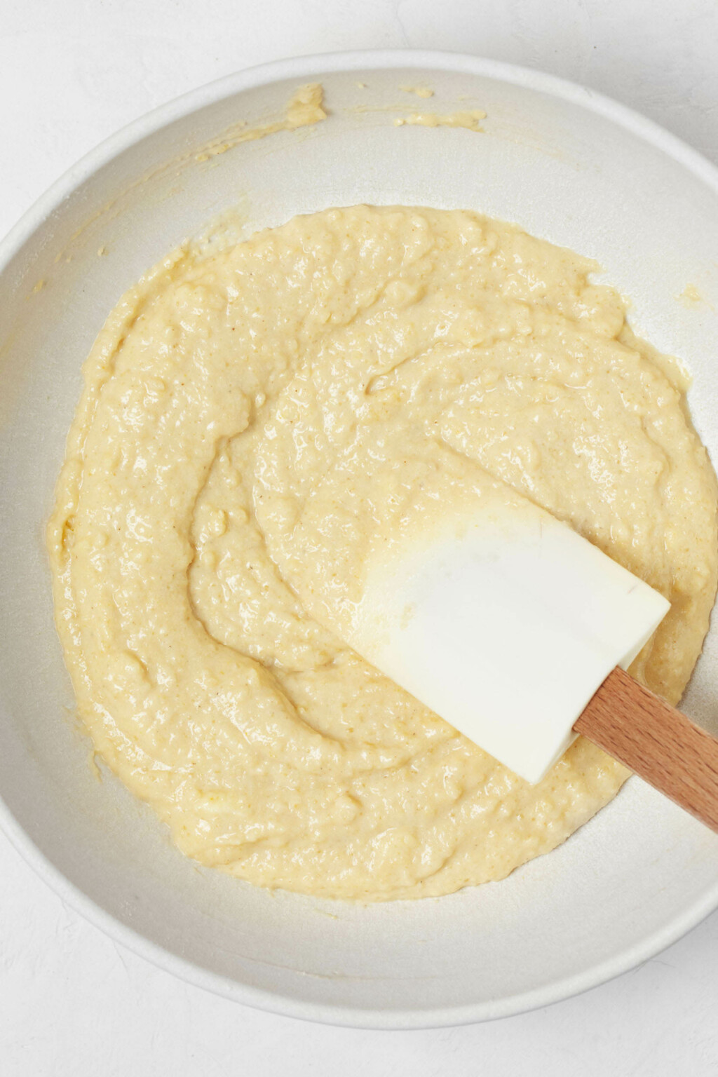 A pale yellow, thick cake batter is being mixed in a white mixing bowl.