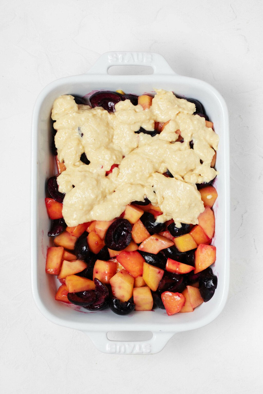 A white, rectangular baking dish has been filled with fresh fruit. It's partially covered with a pale yellow cake batter.