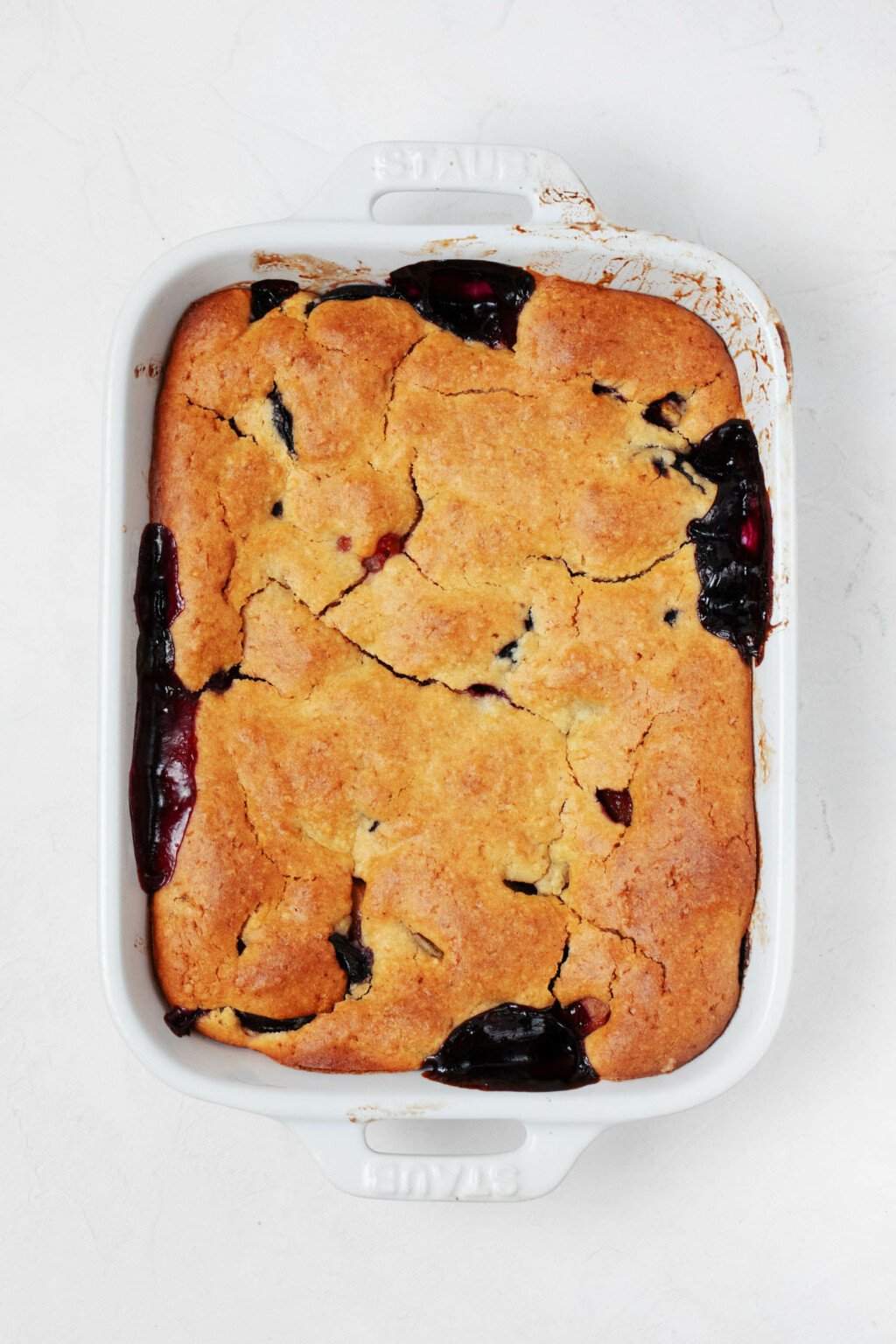 A white, rectangular baking dish holds a vegan peach cherry cobbler with a golden brown, cake-like topping.