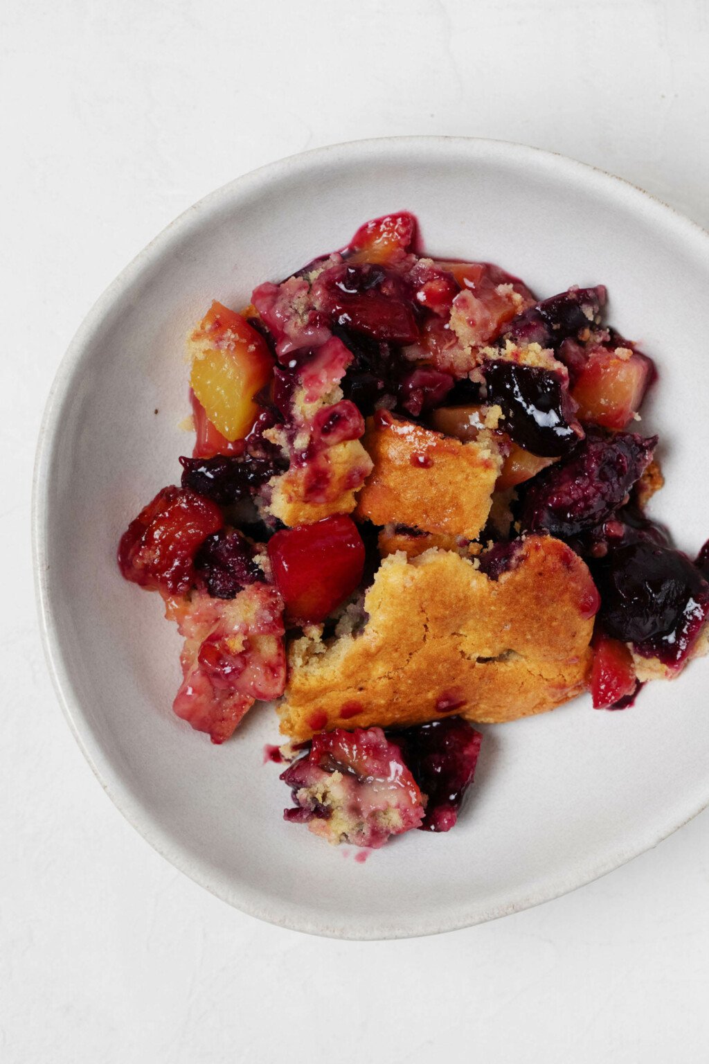 A small, asymmetrical white bowl is filled with peaches, cherries, and a cake-like topping. 
