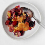 An asymmetrical, white bowl has been filled with a vegan peach cobbler that also has dark red, sweet cherries.