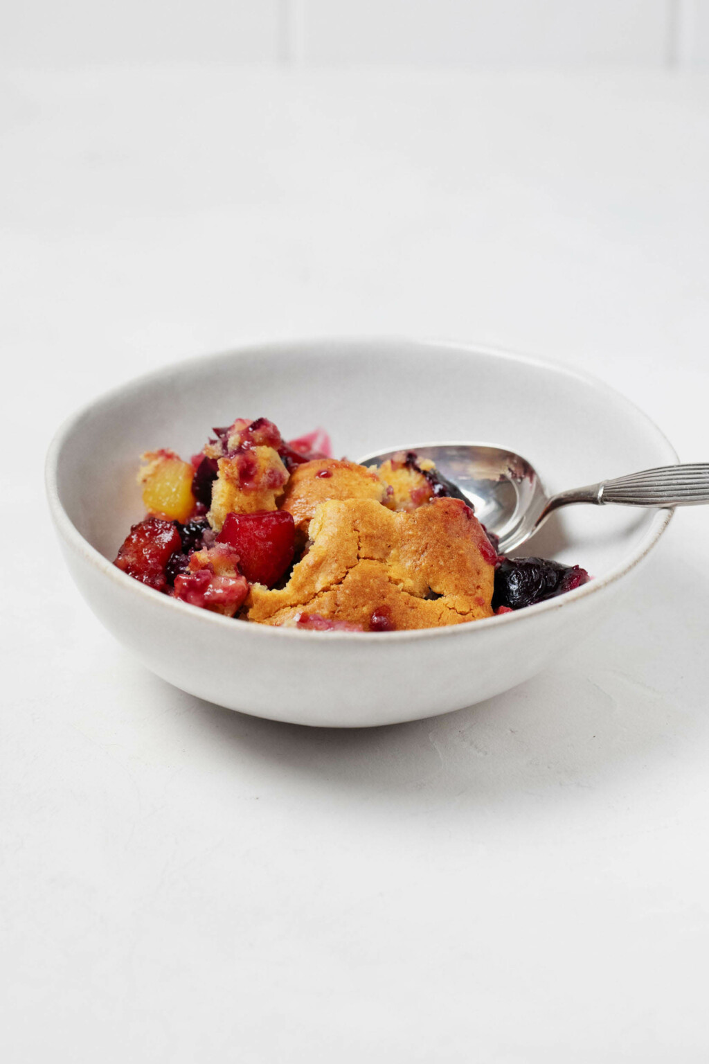 A white, small bowl is filled with a peach and cherry cobbler and soft, cake-like topping. There is a silver spoon resting in the dish, and the dish is on a white surface.