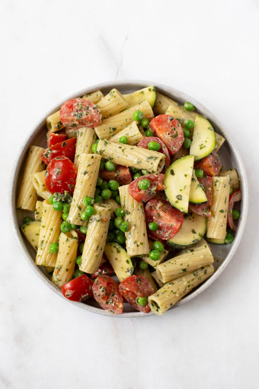 A round, white bowl holds a pasta salad that's made with a tahini base. The pasta salad is full of cherry tomatoes and peas.