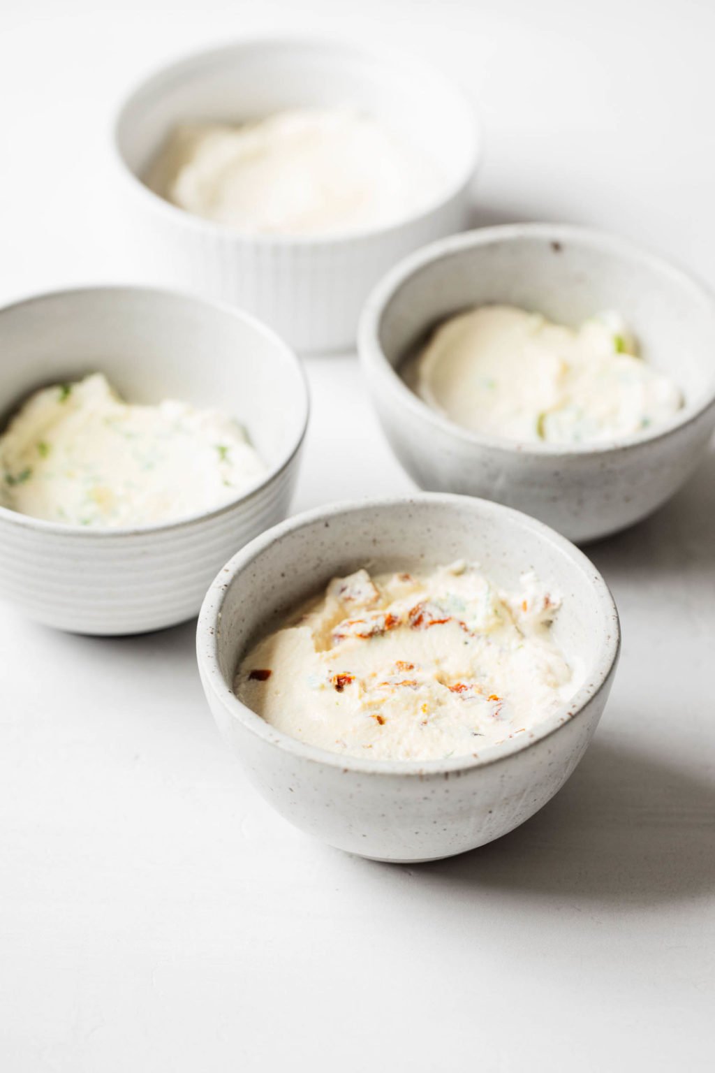 An angled photograph of four small pinch bowls, each filled with a creamy spread, against a white surface.