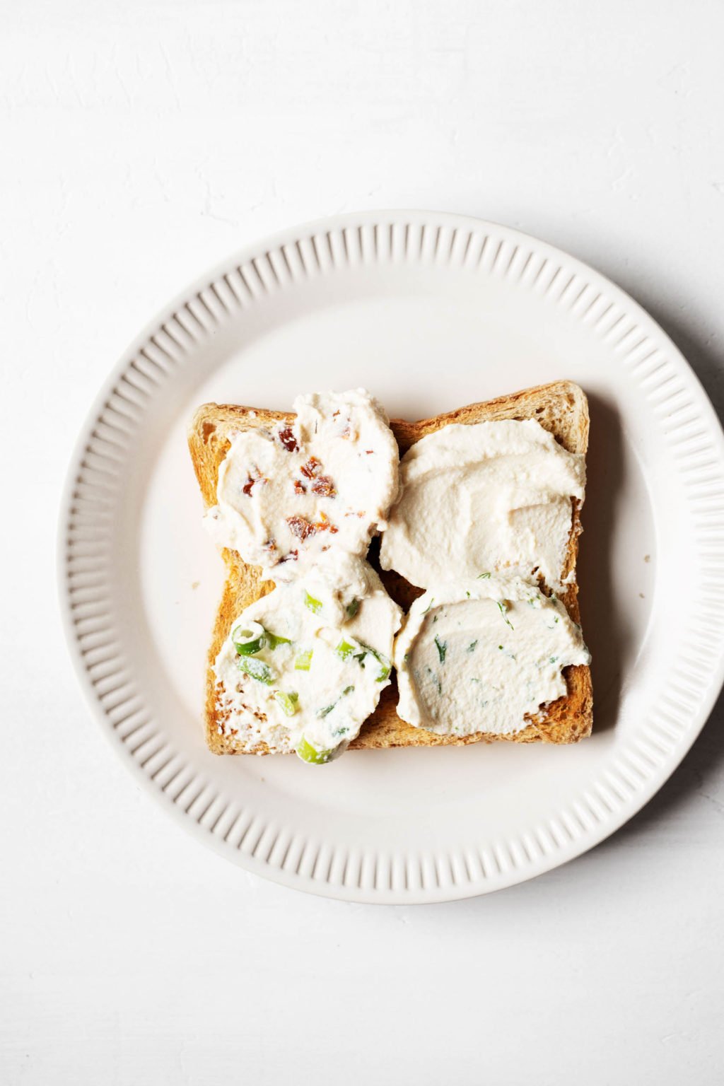 A slice of square toast has been topped with four different varieties of vegan tofu cream cheese. It rests on a fluted white plate.