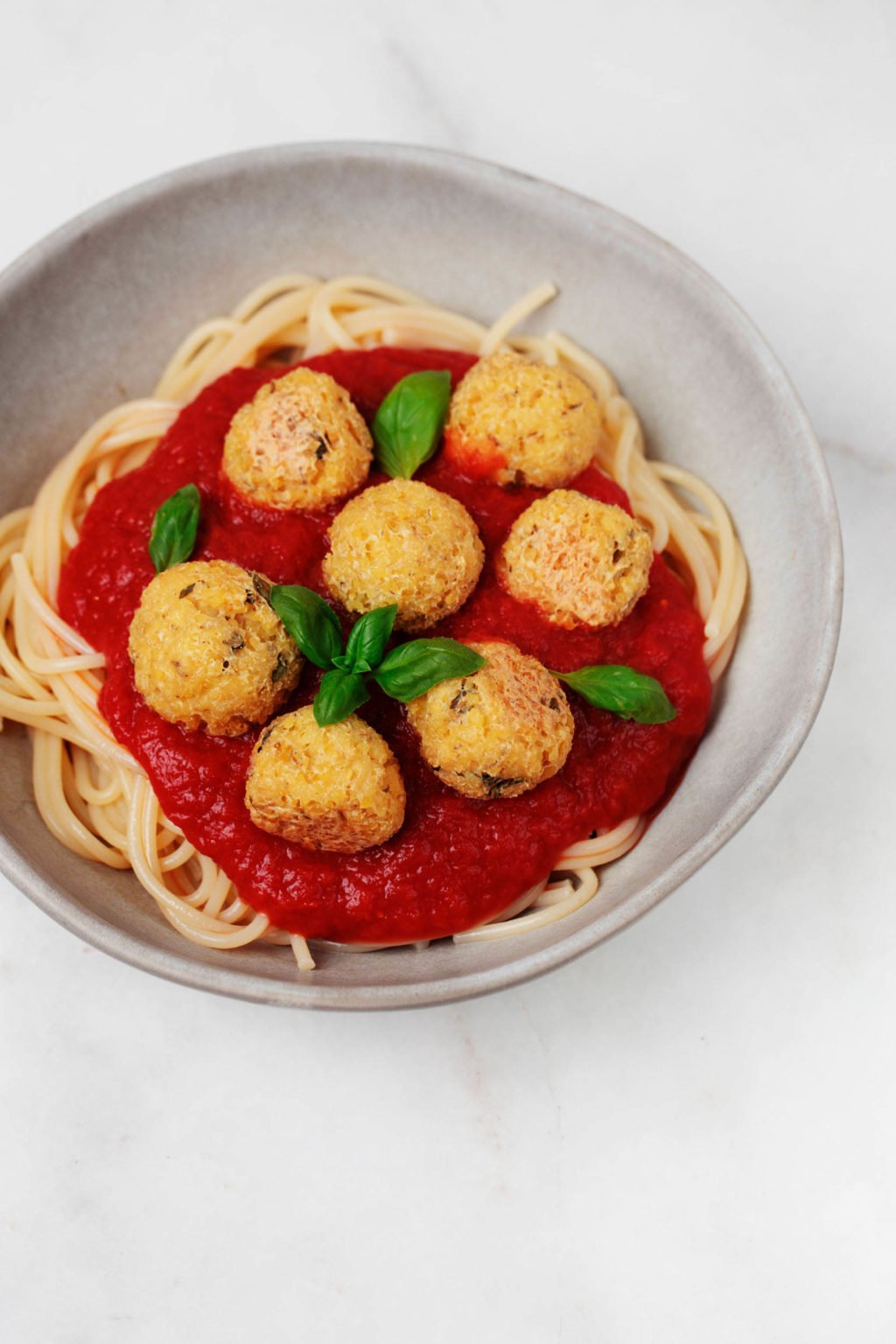A bowl of tofu bulgur meatballs has been smothered by tomato sauce and garnished with green basil leaves.