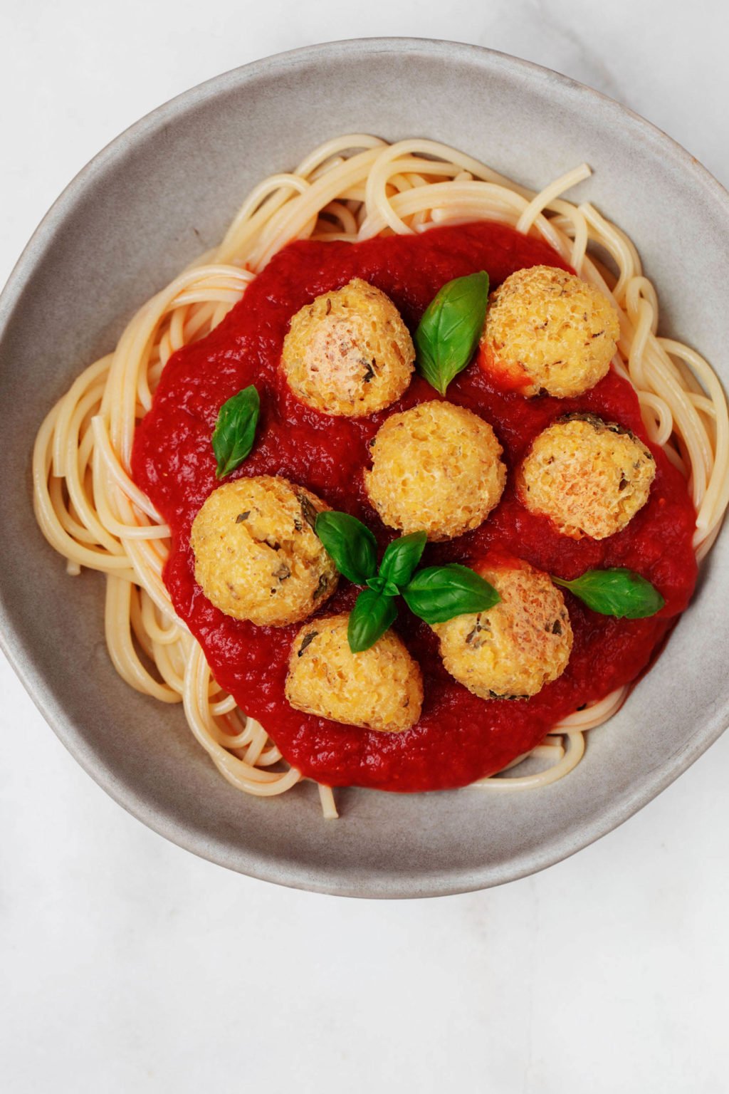 Homemade vegan protein is served over spaghetti and red sauce, in a ceramic bowl. It rests on a white surface.