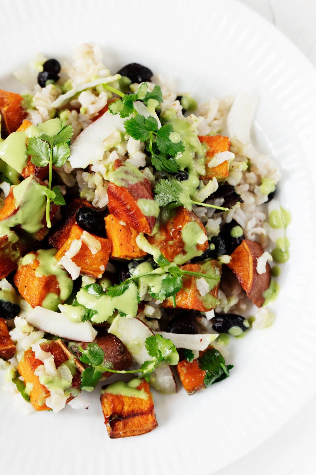 An overhead image of sweet potatoes, brown rice, and black beans. All of the ingredients are mixed with coconut chips and topped with a bright green dressing.