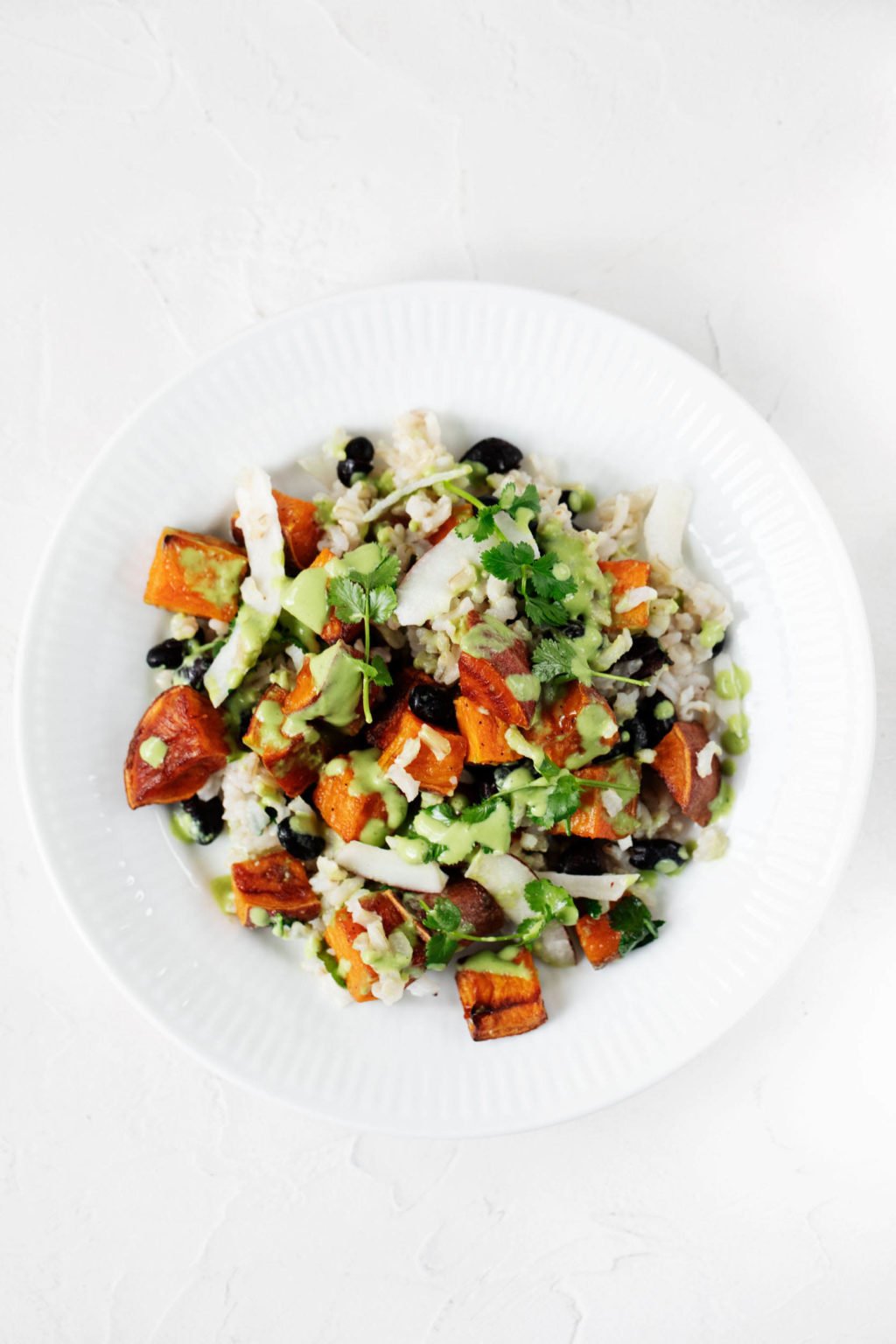 A large-rimmed, white bowl is filled with a mixture of plant-based grains, legumes, and herbs. It rests on a white surface.