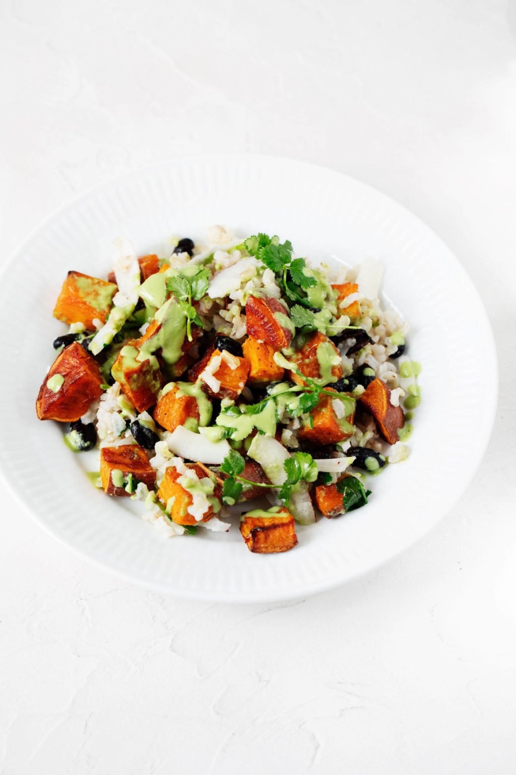 An angled photograph of a wide, white bowl, which has been filled with sweet potatoes, beans, and vegetables. It's topped with a bright green sauce and resting on a white backdrop.