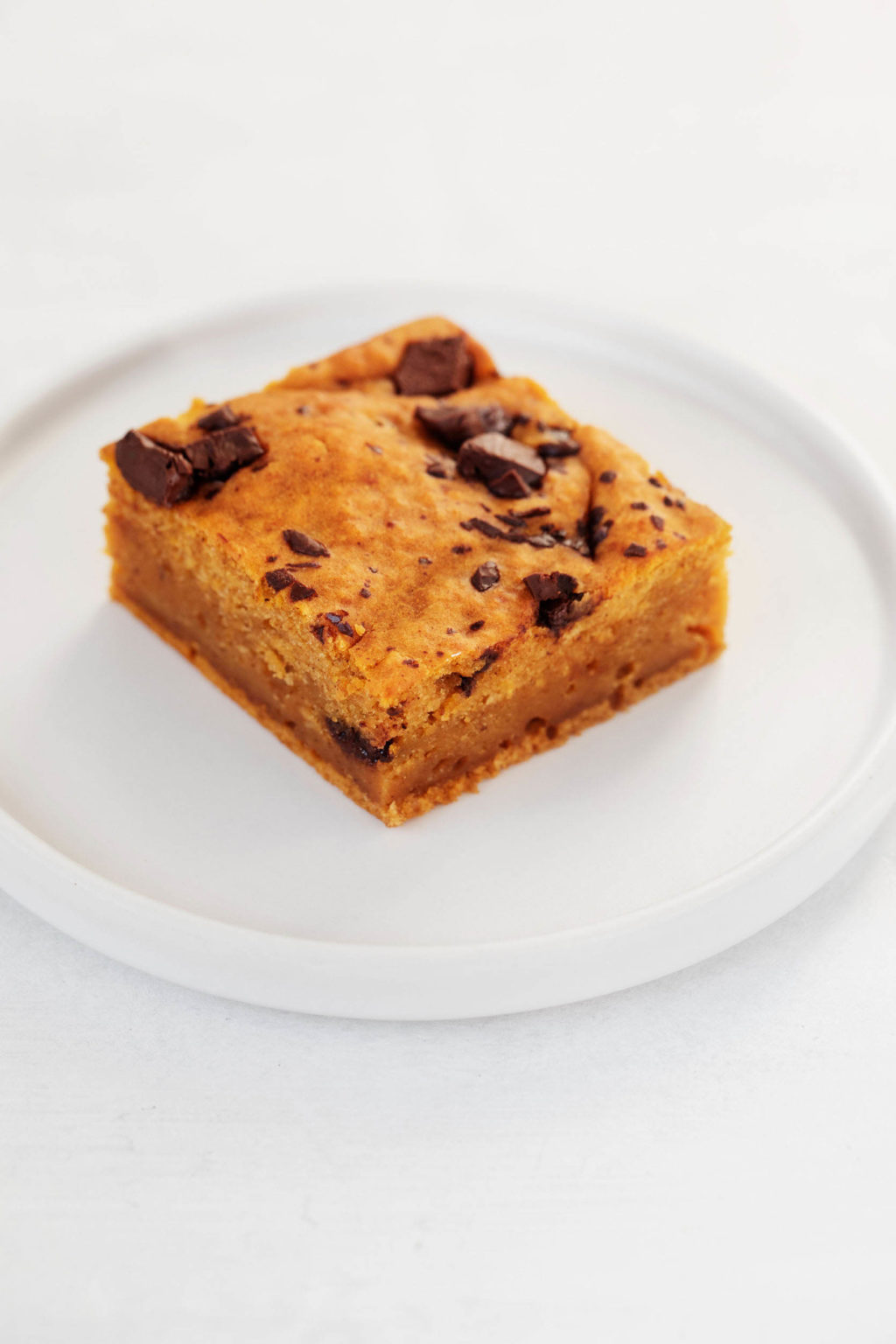 An angled photo of a vegan pumpkin chocolate chip blondie. It's rest on a small, round, white dessert plate.