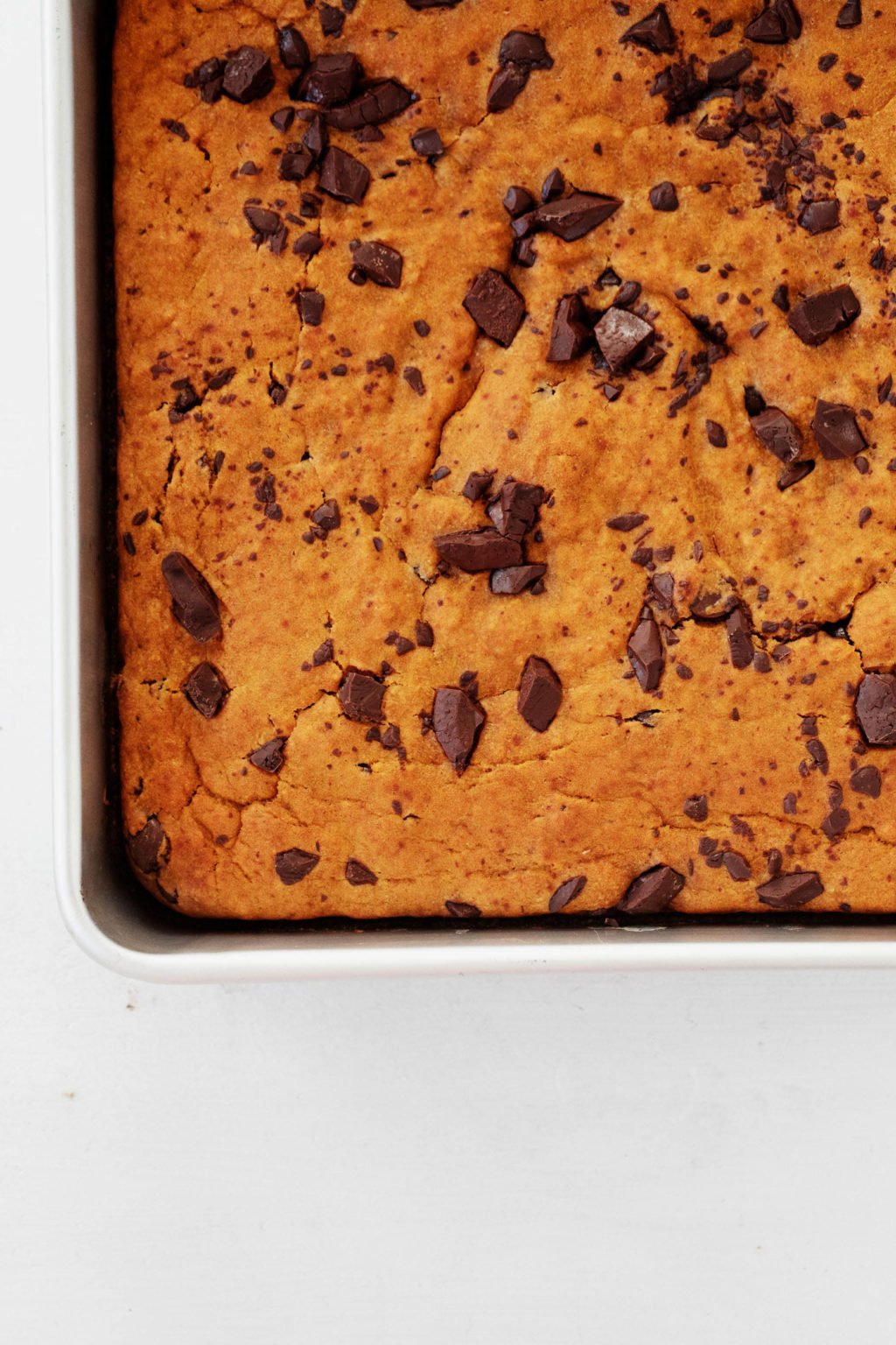 An aluminum baking dish is resting on a white surface. It's filled with a chocolate-studded, plant-based baked good.
