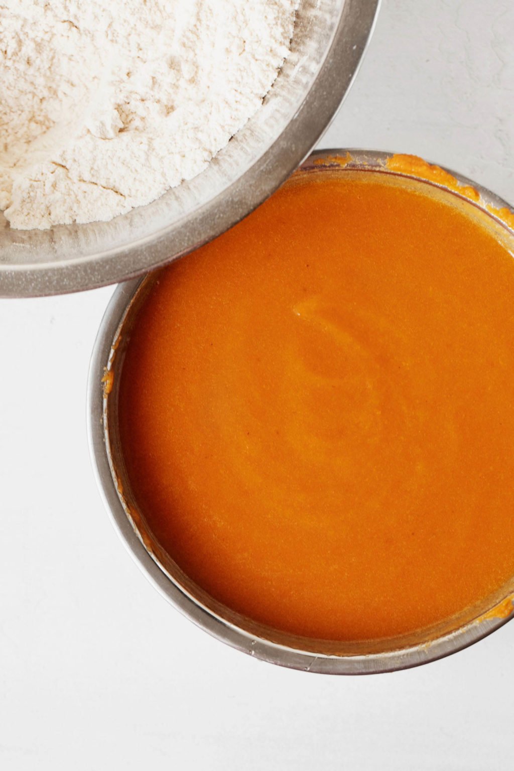 Two mixing bowls. One is filled with flour and one with a wet mixture that includes pumpkin.