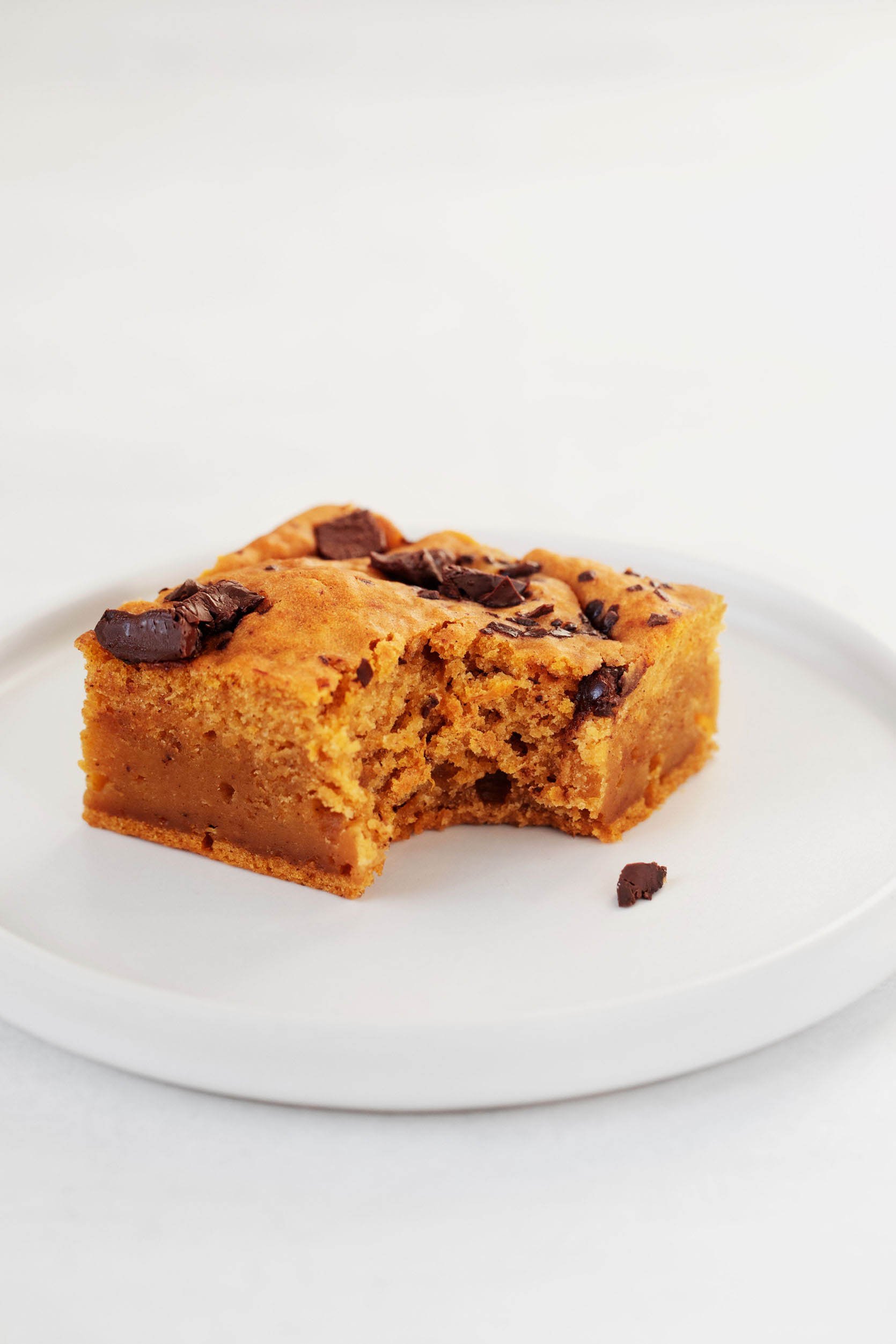 A cakey vegan pumpkin chocolate chip blondie is resting on a small, rimmed, white dessert plate.