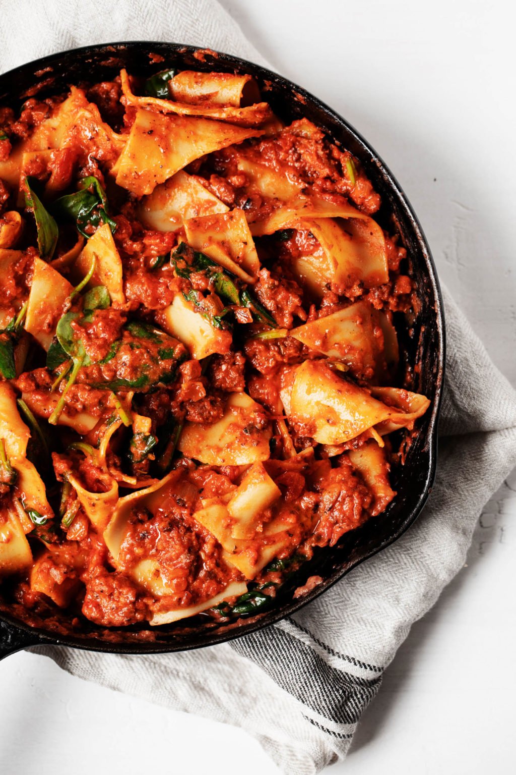 An overhead, close up photograph of a tomato based pasta dish in a large skillet. A cloth napkin is resting underneath the skillet.