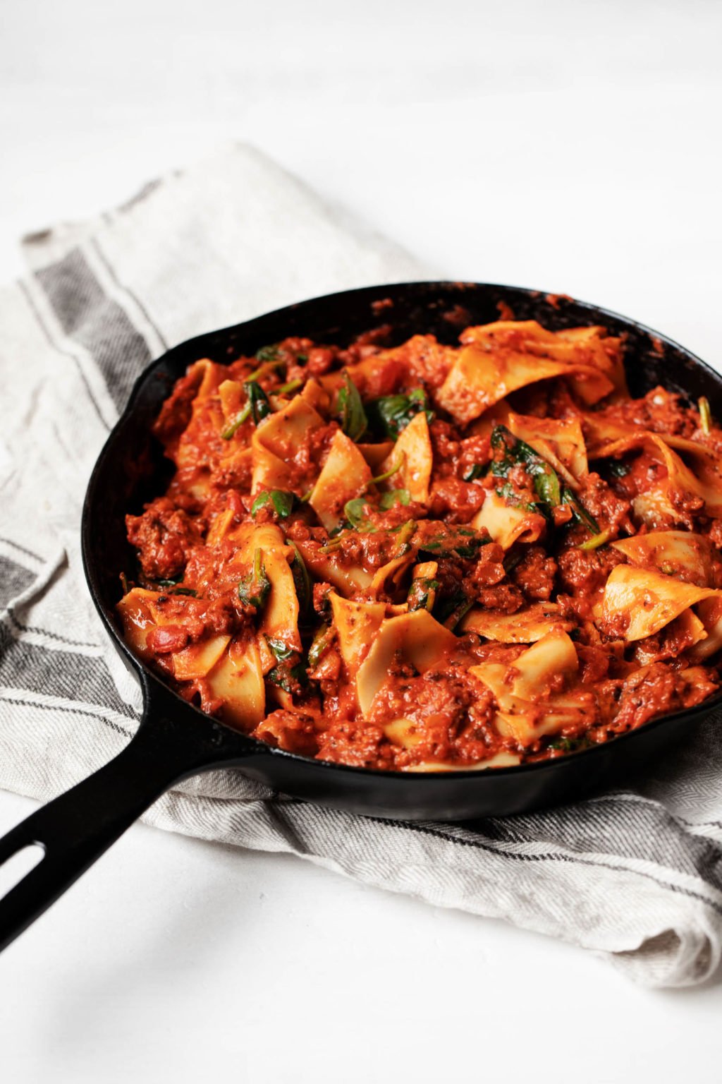 An angled photograph of a black, cast iron skillet, which rests on a striped linen napkin. The skillet is filled with a tomato based pasta dish.