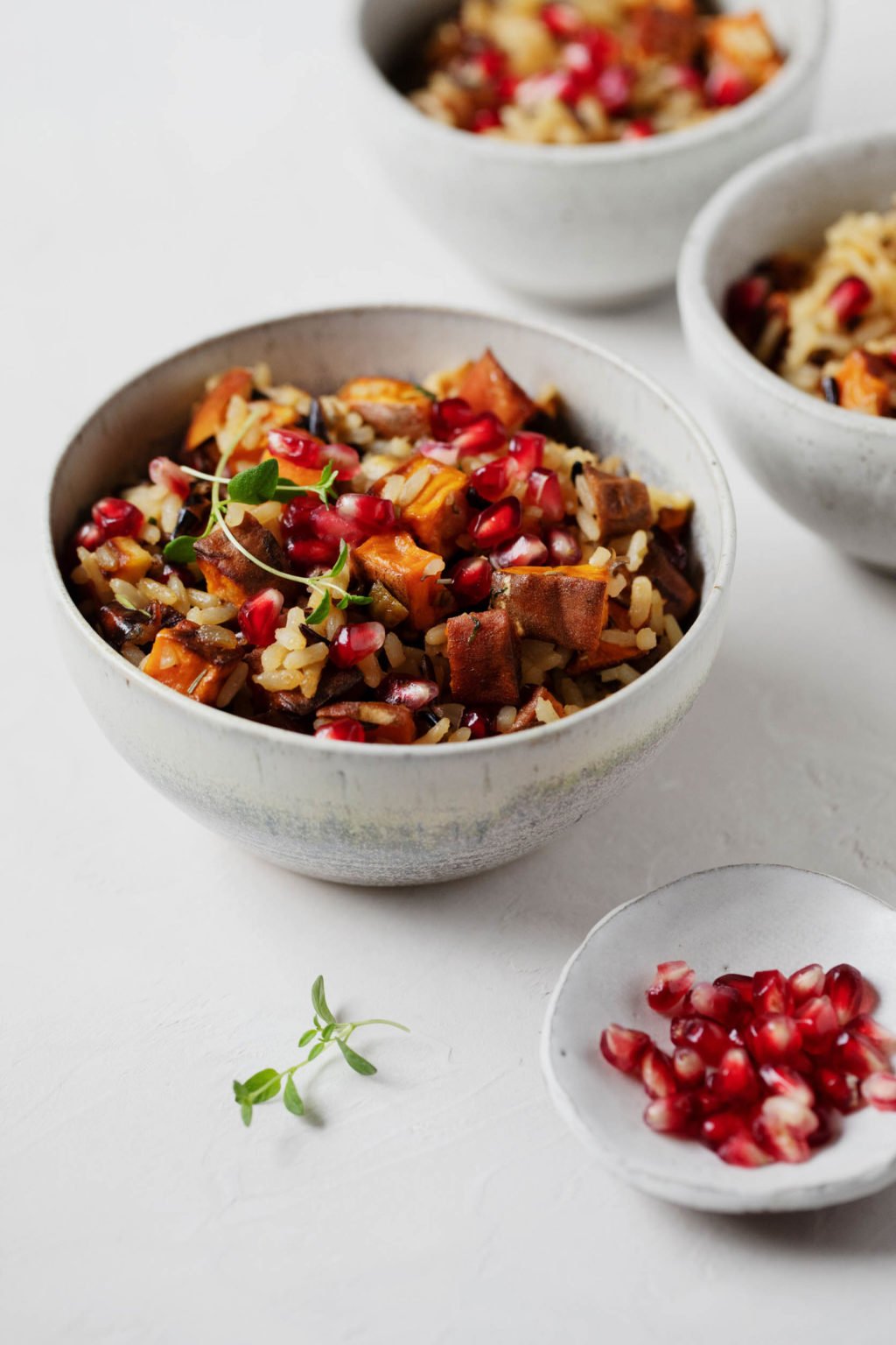 A few bowls of a vegan holiday side dish, with a small pinch bowl of pomegranate arils and fresh herbs nearby.