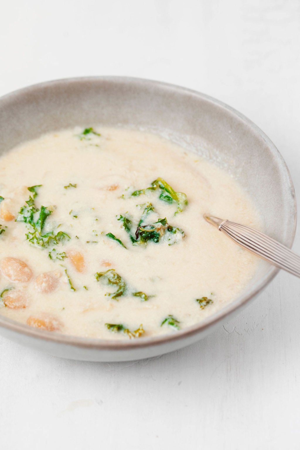 A ceramic bowl has been filled with a cream-colored soup and leafy greens. It's resting on a white surface.