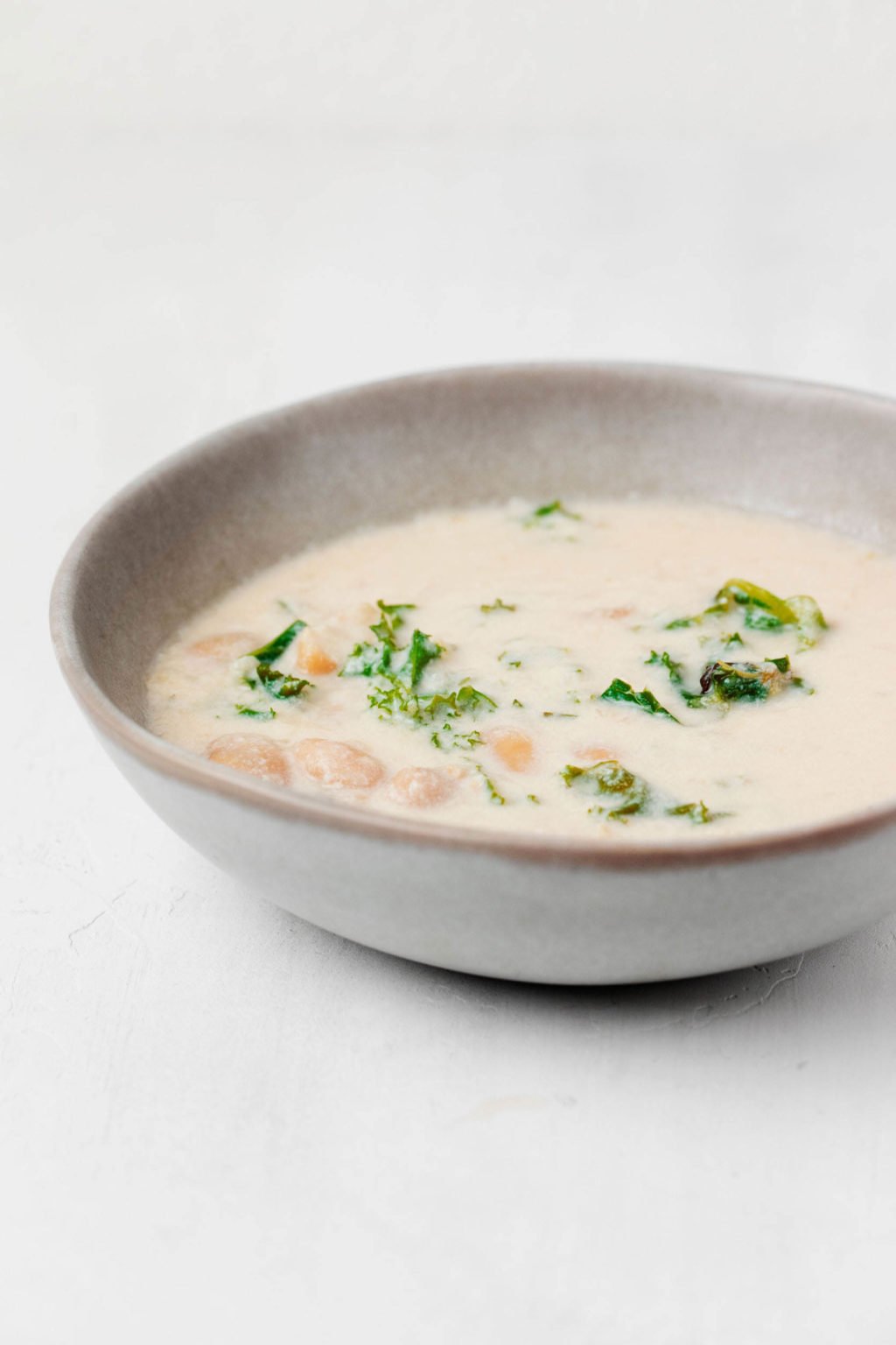 An angled photograph of a light gray, ceramic bowl. It's filled with a creamy mixture of legumes and greens. It rests on a white surface.