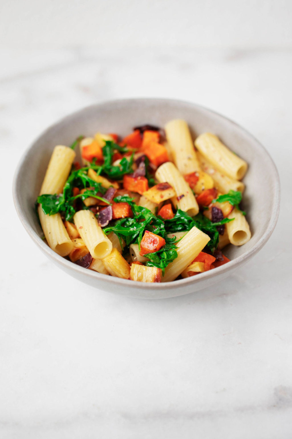 A gray bowl, filled with colorful vegetables and pasta, rests on a white marble surface.