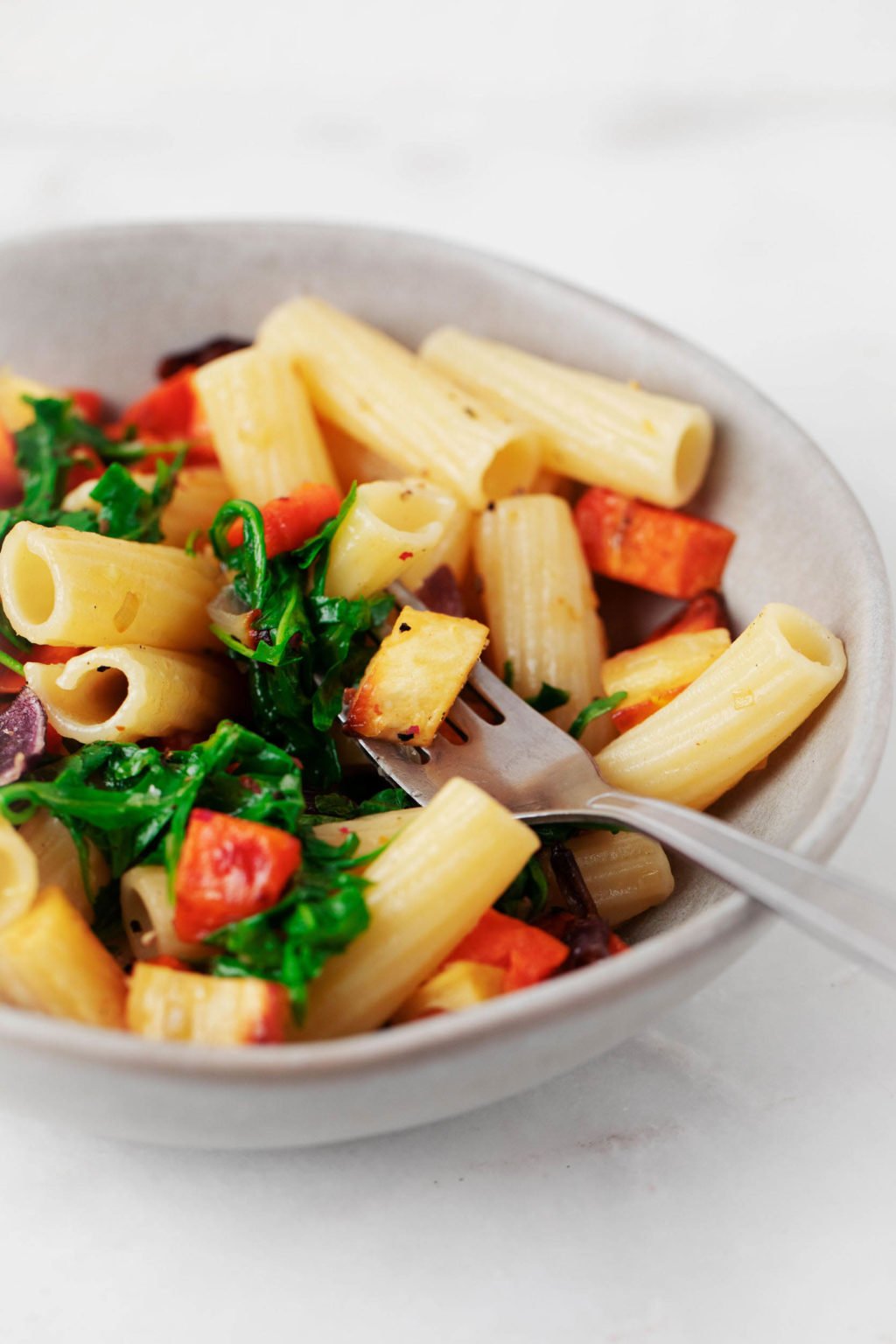 A gray ceramic bowl is filled with a balsamic roasted vegetable pasta. There's a fork piercing a piece of the pasta.