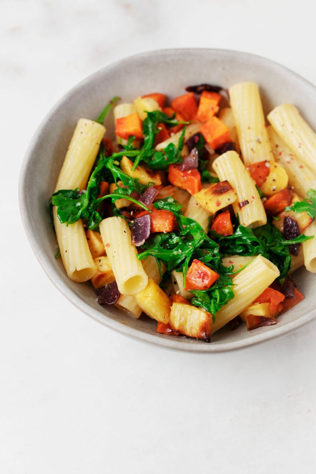 A colorful mix of rigatoni, arugula, and carrots is served in a gray ceramic bowl.