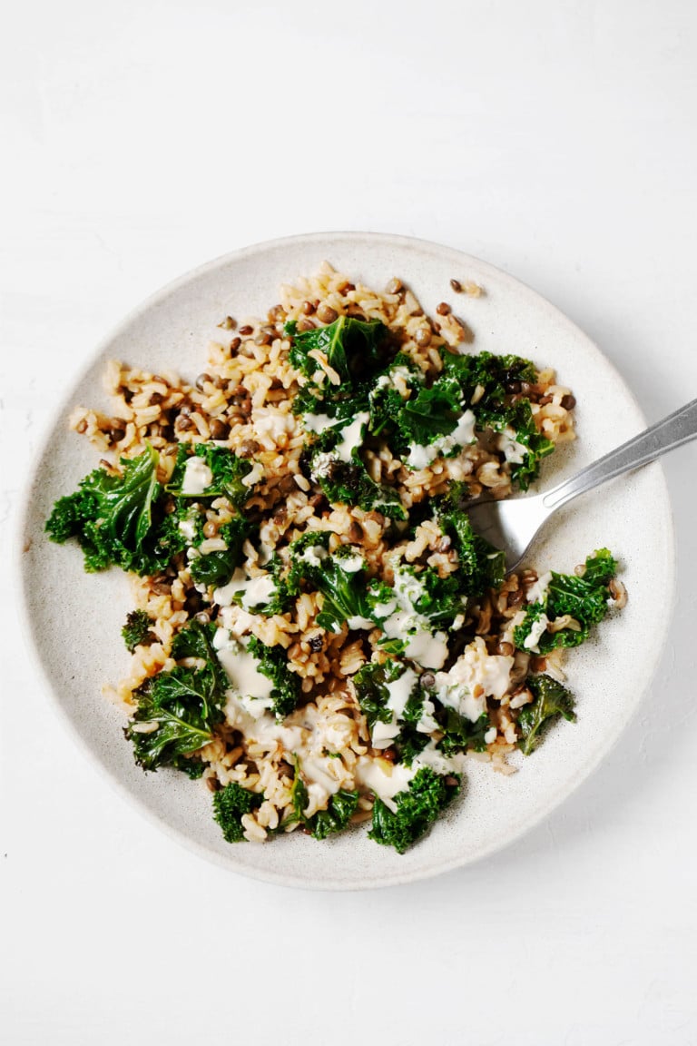 A round, white plate is covered with cumin-spiced lentils & rice, a serving fork, and a drizzle of pale-colored tahini sauce.