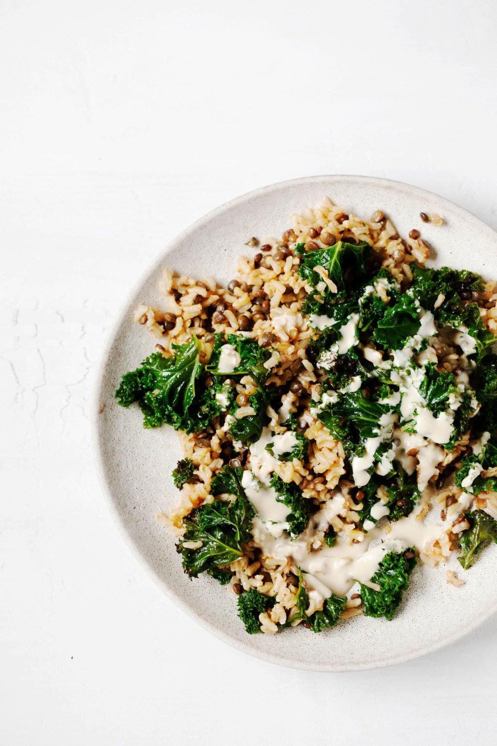A pale, ceramic plate has been topped with a vegan dish of rice, lentils, kale, and a creamy sauce.