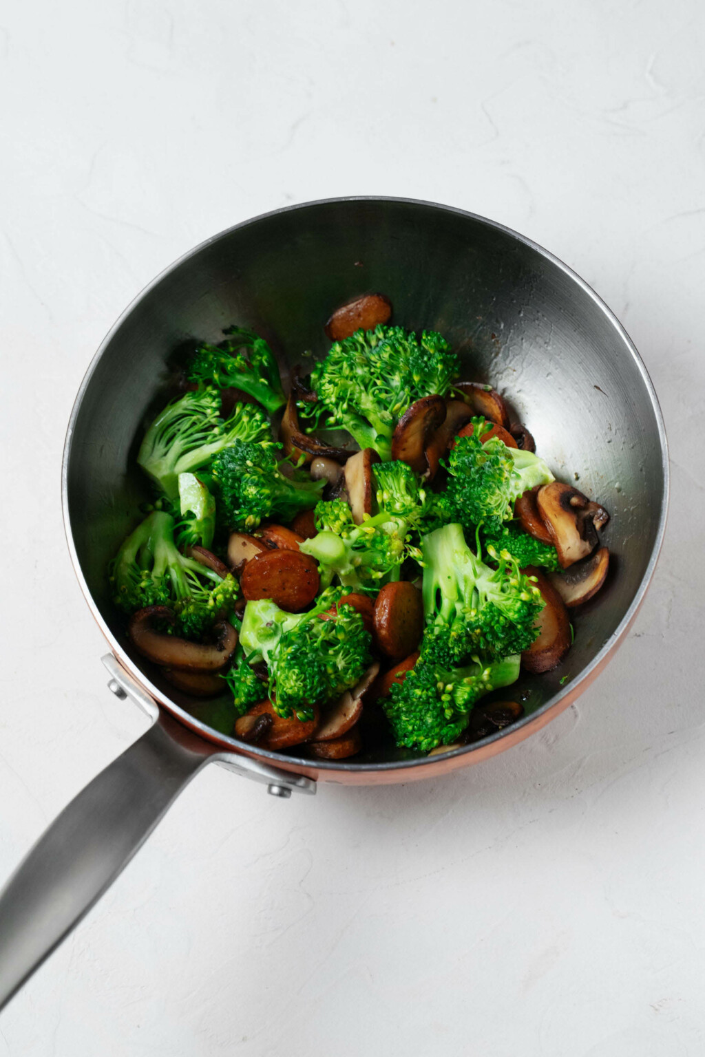 A silver skillet is being used to sauté sliced mushrooms and sausage, along with bright green broccoli florets.