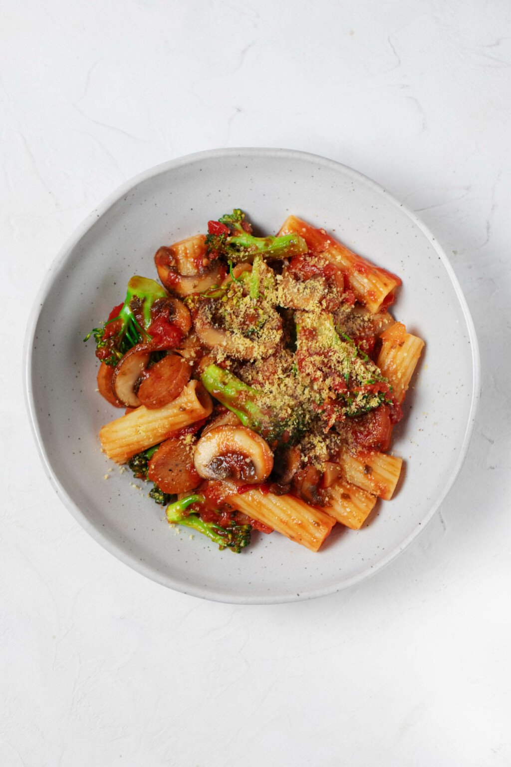 An overhead image of a white bowl, which is filled with a vegan pasta dish made with plant-based sausage, broccoli, and marinara sauce.