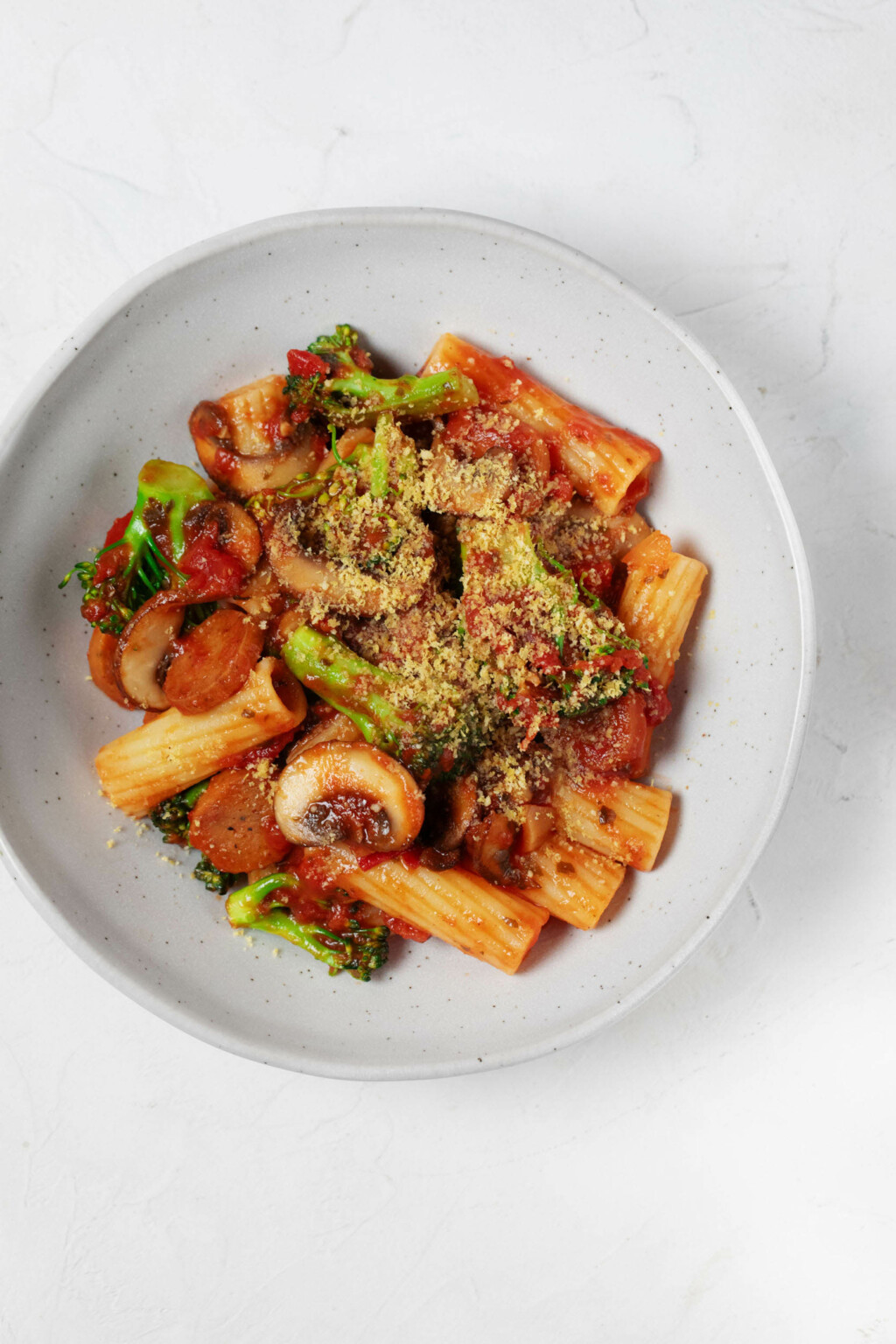 An overhead image of a white bowl, which is filled with a vegan pasta dish made with plant-based sausage, broccoli, and marinara sauce.